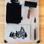 Top-down view of a wooden desk showing black ink rolled out into a rectangle on a glass inking plate, an ink-covered brayer/roller, a palette knife, and the carved linoleum block of penguins.