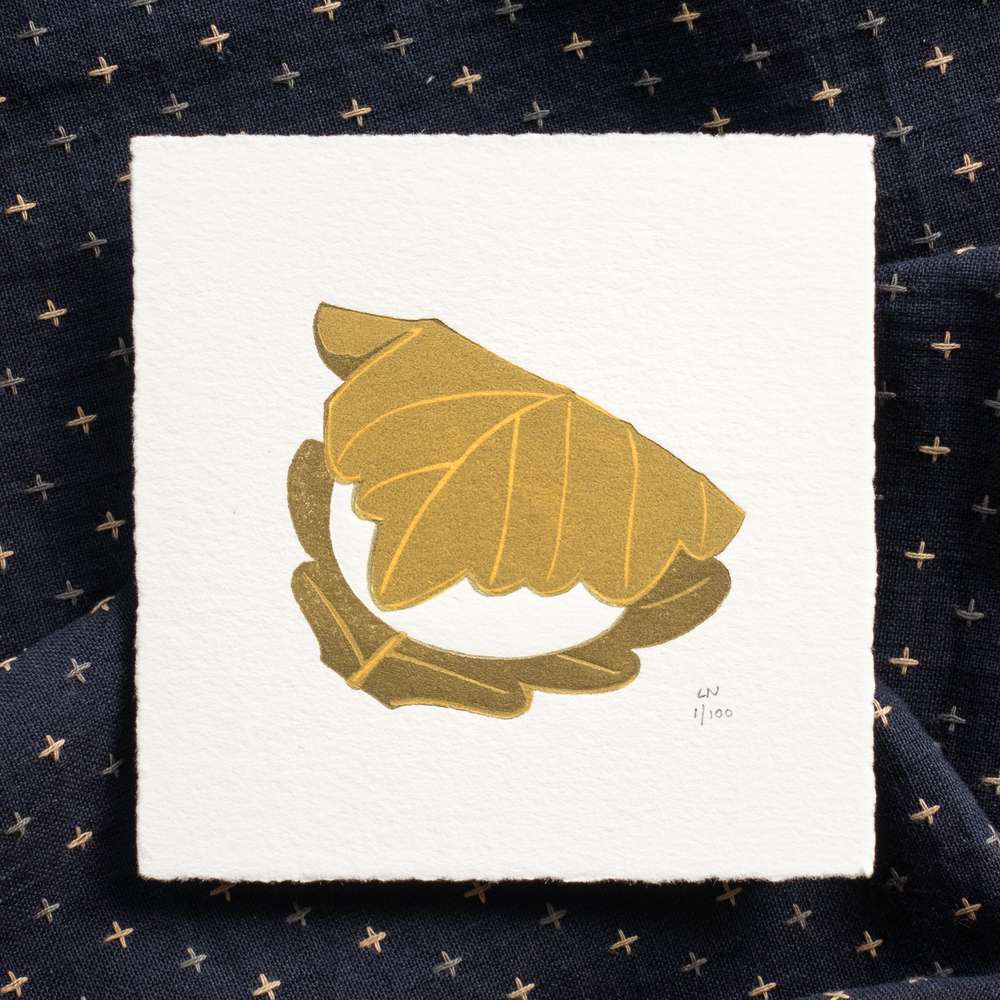 A square piece of paper depicting a kashiwa mochi - a white ball wrapped in an olive green oak leaf. The print is signed in the bottom right corner. The print sits on crumpled dark navy fabric with light-colored checks.