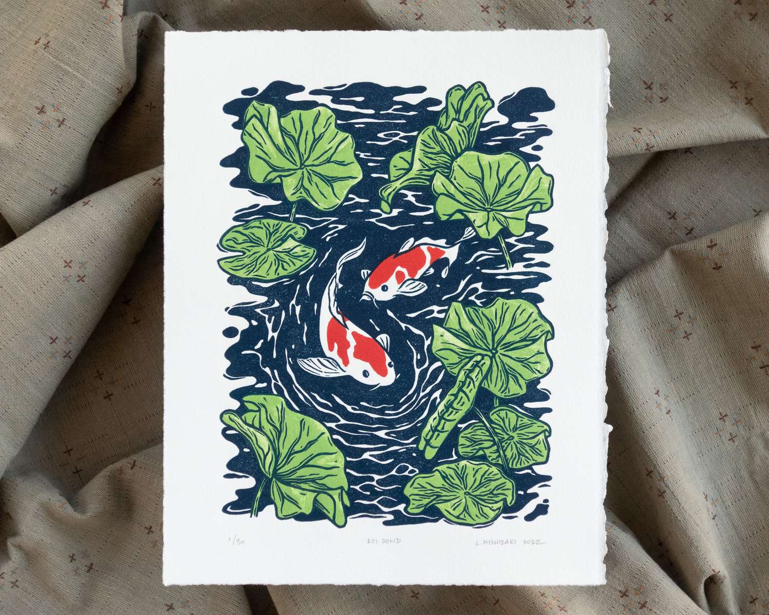 A white vertical rectangular paper depicting two red and white koi swimming below the rippled surface of a pond, with many green lily pads growing out of the water. The print sits on top of a rumpled grey fabric backdrop.