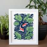 The blue, green, and red print depicting koi swimming in a lotus pond, framed in a white frame and sitting on a brown table with house plants.