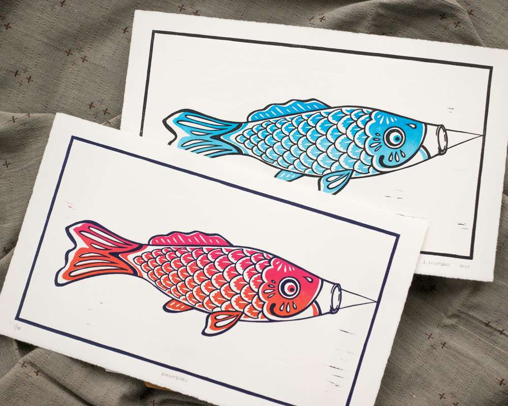 Two vibrantly colored koinobori prints on white paper, overlapping. The top print is a blue fish; the bottom print is an orange/pink fish.