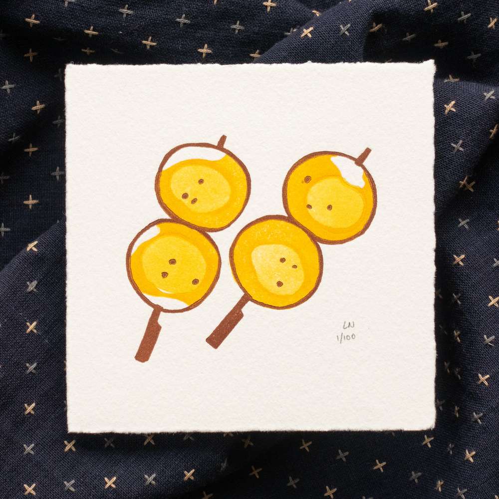 A square print on top of rumpled navy fabric with light colored X's. The print is done in three colors and depicts two brown skewers each containing two mochi balls, covered in a yellow-orange sauce. The print is signed in the bottom right corner with 'LN 1/100'.