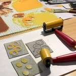 Two red-handled brayers inked in yellow/gold ink sit on a table. Next to them are three carved linoleum blocks, inked in brown, yellow, and golden yellow; these three blocks form the mitarashi dango print design. Other tools and a glass plate containing rolled-out paint swatches sits in the background.