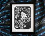 Black and white blockprint of a monkey in a persimmon tree, framed in a thin black frame. The frame sits on top of rumpled blue striped fabric.