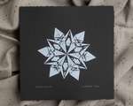 A white impression of an origami snowflake on square black paper.