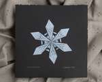 A white impression of an origami snowflake on square black paper.