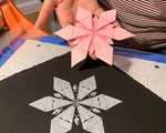 A white impression of an origami snowflake on black paper, and a hand holding the origami snowflake.