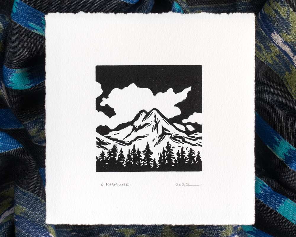 A white square print depicting fluffy clouds, a pointy double mountain peak, and conifers in stark black. The paper sits above rumpled blue and black striped fabric.