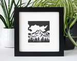 A black and white print of a mountain and trees, inside a square black frame in front of two potted house plants.