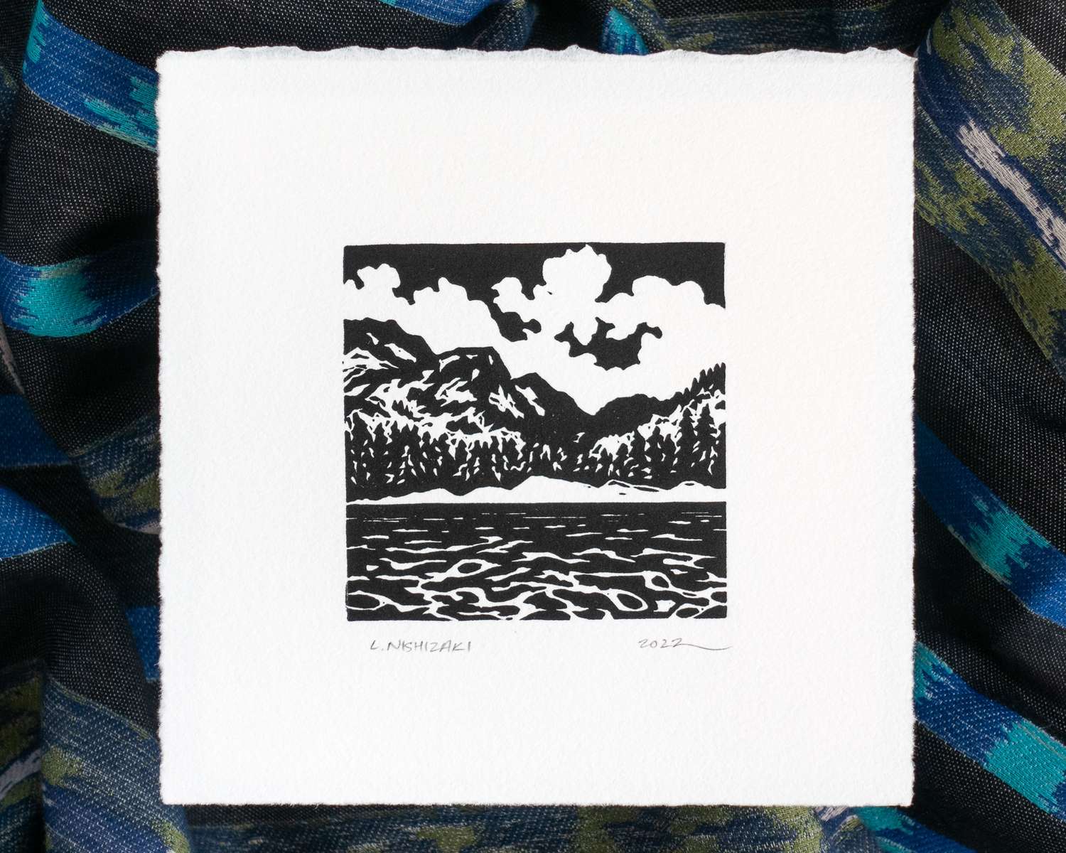 A square white print depicting clouds, a ragged ridgeline, trees, and the surface of a lake, using black. The paper sits above rumpled blue and black fabric.