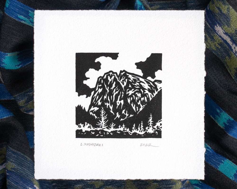 A highly textured black and white depiction of Mt. Index with clouds behind the mountain and trees in the foreground. The design is depicted in black ink on white paper, and the paper sits above dark rumpled fabric.