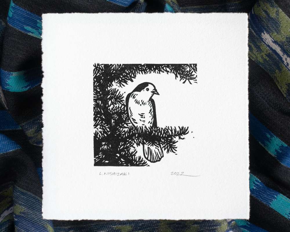 A black-and-white print of a canada jay sitting on a tree branch. The print is on white paper that sits above dark rumpled fabric.