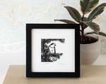 A framed black and white print of a bird, sitting on a wood shelf in front of a house plant.