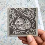 A right hand holds a carved grey linoleum block above a paper map of the wilderness.