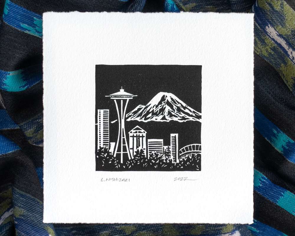 A black and white print depicting the Space Needle and downtown Seattle skyline, with Mt Rainier rising in the distance. The print is on white paper, and sits above dark rumpled fabric.