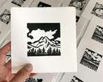 A left hand holds up an unsigned square print of a mountain, with out-of-focus black and white prints in the background.