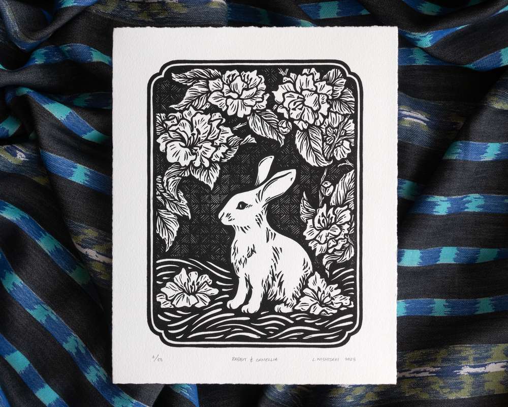 A black and white blockprint of a rabbit underneath a camellia bush with blossoms. The edges of the paper are hand-torn, and the print sits on top of rumpled blue striped fabric.