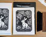 A stack of signed rabbit blockprints and various carving paraphenalia, arranged on a wooded desk.