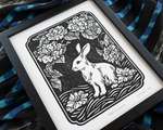 A black and white illustration of a rabbit and camellia blossoms, lying on top of a rumpled blue striped piece of fabric.