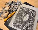 A messy desk covered with a carved linoleum block, carving tools, and linoleum shavings of various sizes.