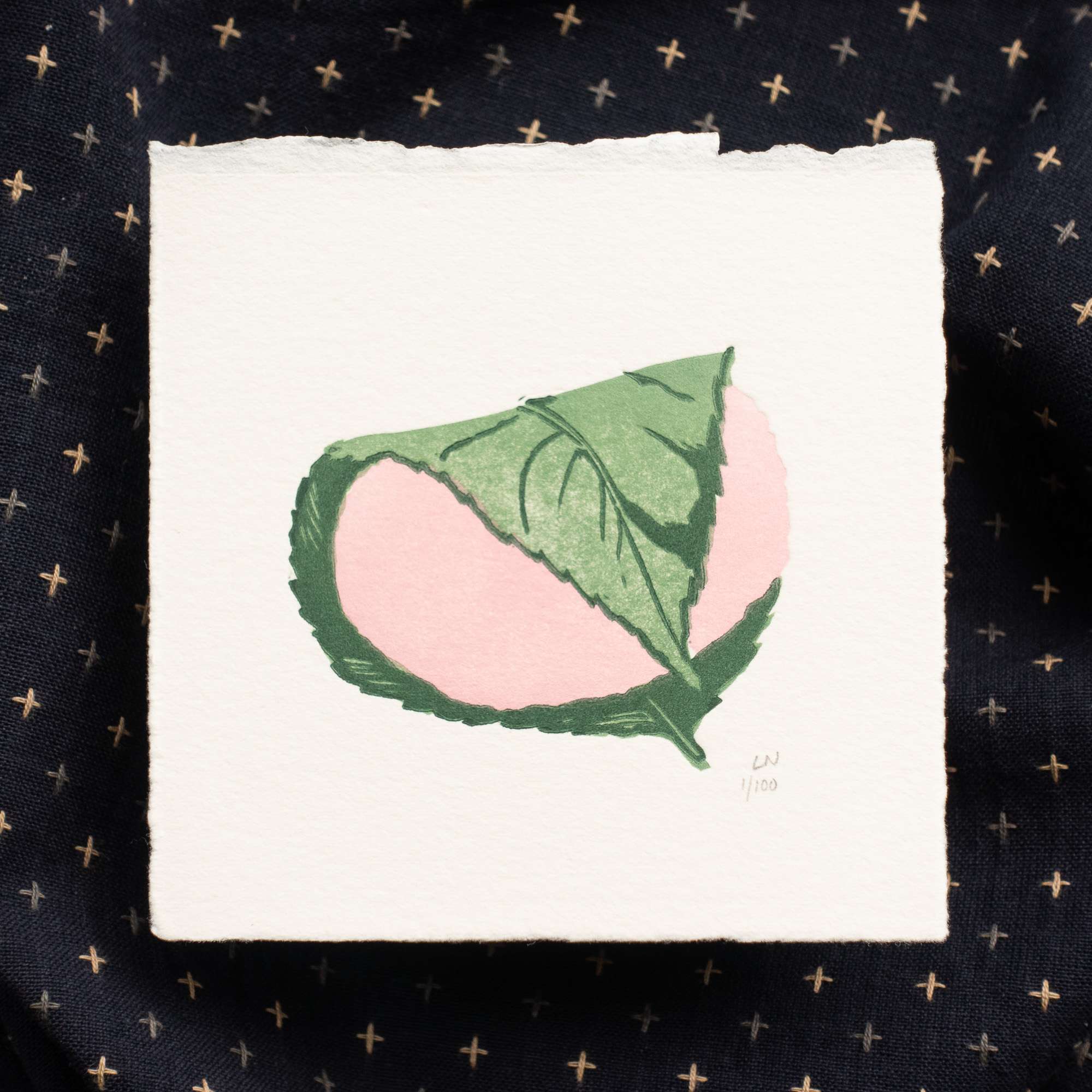 A pink and green print of a Japanese mochi dessert, printed using simple block shapes on thick cream colored paper. The print is labelled on the bottom right edge with the artist initials LN and the edition number 1/100. The paper has rough textured edges (the top edge of the print is a deckle edge). The print lies on top of a geometric navy piece of fabric.