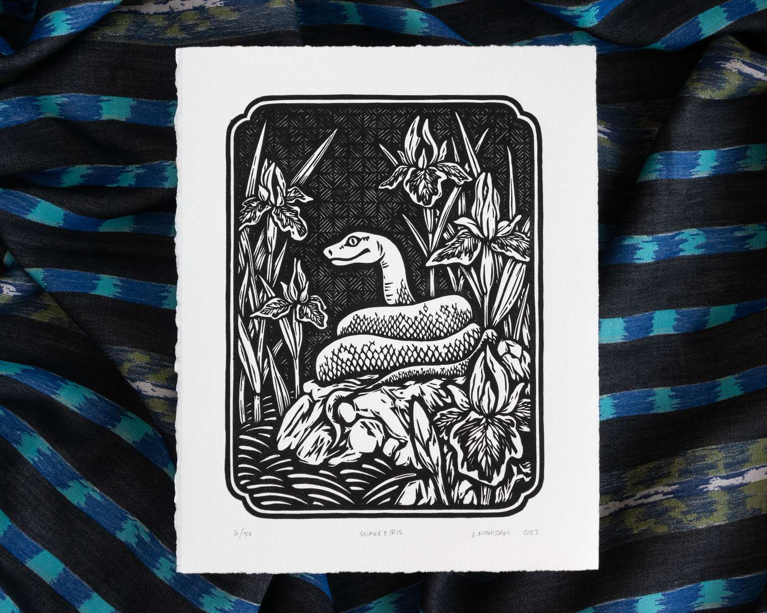 A black and white blockprint of a snake sitting coiled up on a rock, surrounded by irises in bloom. The edges of the paper are deckled and hand-torn, and the print sits on top of a backdrop of rumpled striped fabric.