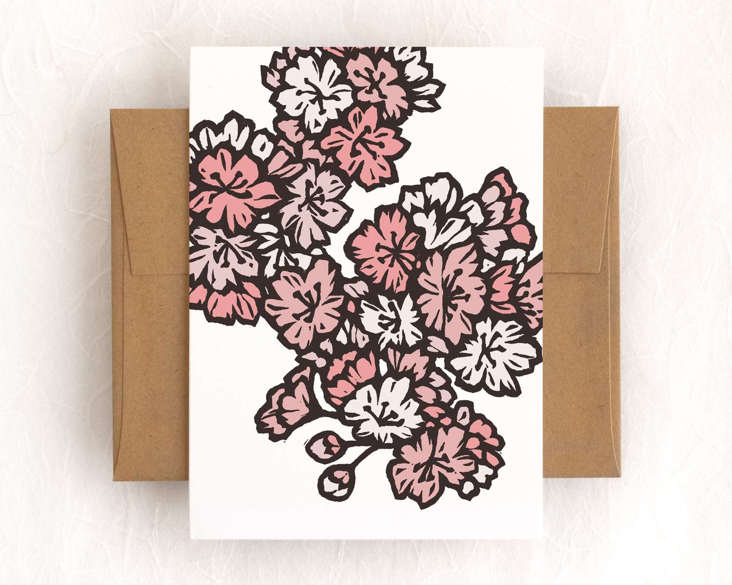 A vertical white card featuring pink blockprinted cherry blossoms. The card sits on top of a brown envelope, which lies on top of a white backdrop.