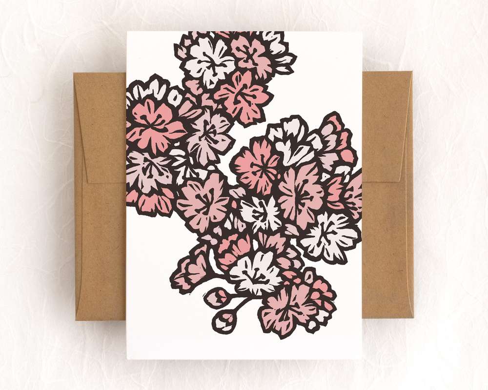 A vertical white card featuring pink blockprinted cherry blossoms. The card sits on top of a brown envelope, which lies on top of a white backdrop.