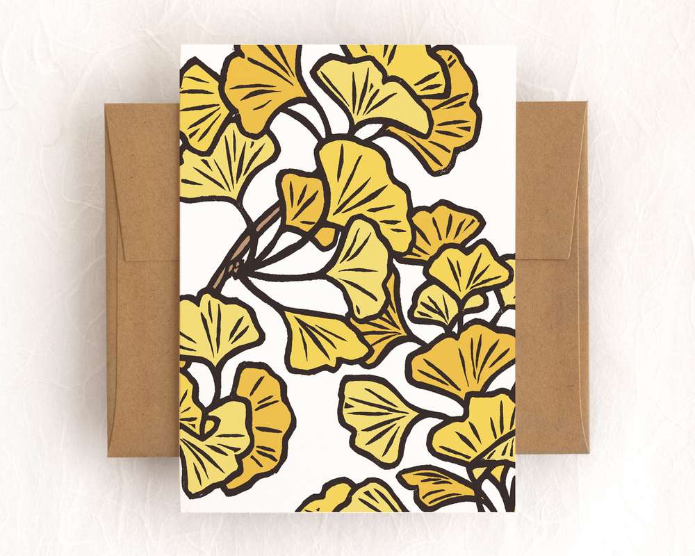 A vertical white card featuring yellow ginkgo leaves on branches. The card sits on top of a brown envelope, which lies on top of a white backdrop.