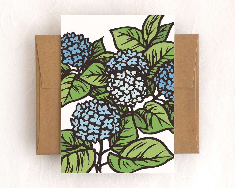 A vertical white card featuring blue blockprinted hydrangea flowers and green leaves. The card sits on top of a brown envelope, which lies on top of a white backdrop.