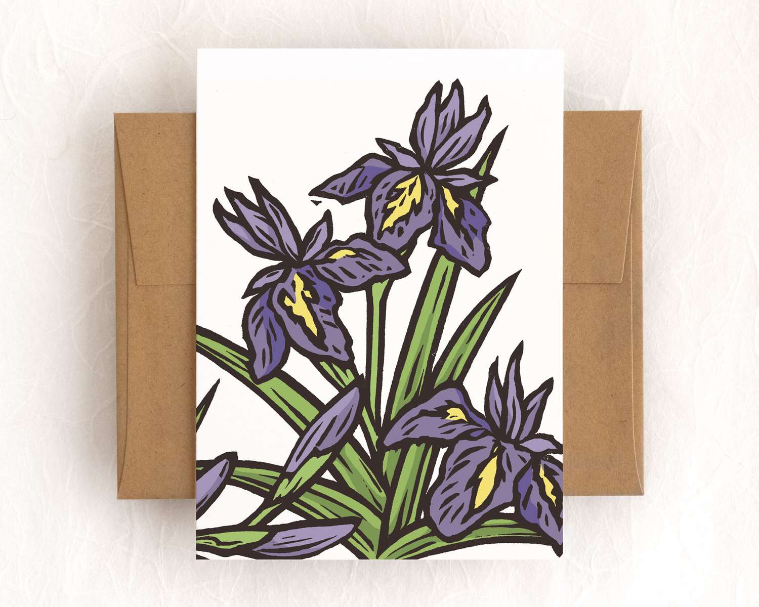 A vertical white card featuring a purple blockprinted iris flowers. The card sits on top of a brown envelope, which lies on top of a white backdrop.