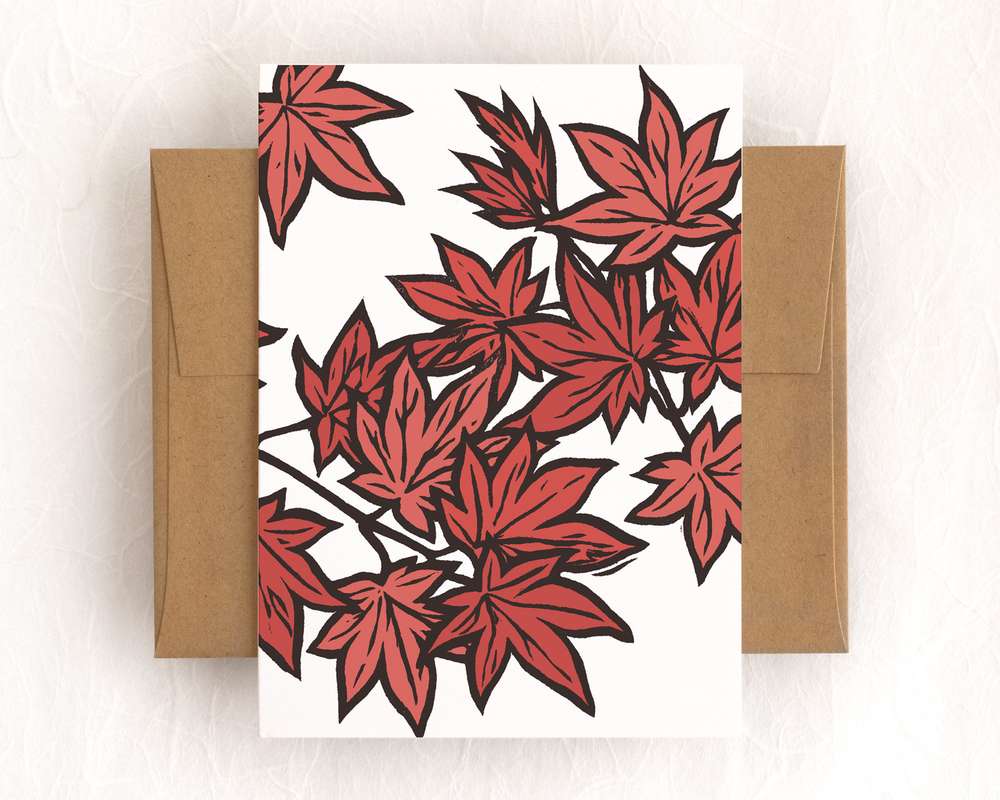A vertical white card featuring red japanese maple a orange persimmon and green leaves on branches. The card sits on top of a brown envelope, which lies on top of a white backdrop.