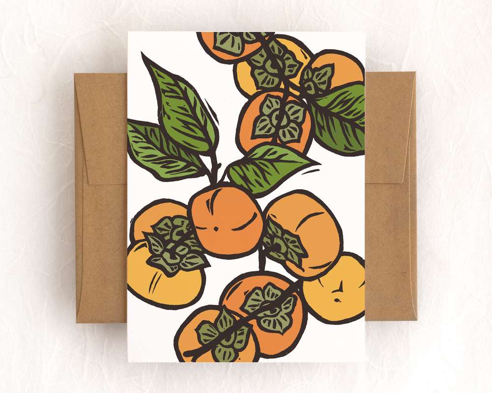 A vertical white card featuring a orange persimmon and green leaves on branches. The card sits on top of a brown envelope, which lies on top of a white backdrop.