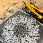 A half carved grey linocut block of a sunflower. The design is darker than the surrounding; the negative space has been carved away. Curled bits of linoleum litter the desk and 2 yellow-handled carving tools sit above the block.