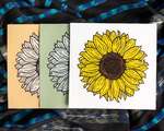 Three overlapping square prints of sunflowers. The right-most is yellow and brown with black outlines, printed on white paper. The middle and left are printed in white ink with black lines on green and pale brown paper, respectively. The prints are on top of rumpled black and blue striped fabric.
