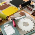 Desk surface covered in printing tools, including multiple brayers and a glass inking plate covered in yellow, black, white, and brown ink, and two carved linoleum blocks covered in white and brown ink used to print sunflowers.