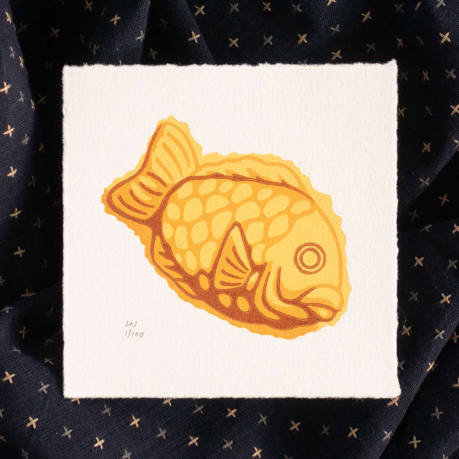 A square piece of paper depicting a taiyaki - a yellow/orange fish shape seen side-on, looking to the right. The print is signed in the bottom left corner. The print sits on crumpled navy fabric with light-colored checks.