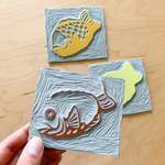 A left hand holds a flat piece of linoleum carved with the dark outline of a taiyaki, covered in dark brown ink. Two more carved linoleum blocks sit in the background, covered in orange and yellow inks.