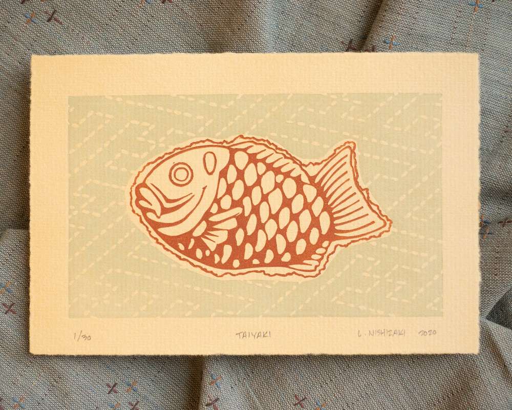 A single taiyaki print on light tan paper, depicted in brown with a pale blue grey background decorated with a sashiko stitch pattern. The print sits on top of an artfully crumpled grey piece of cloth.