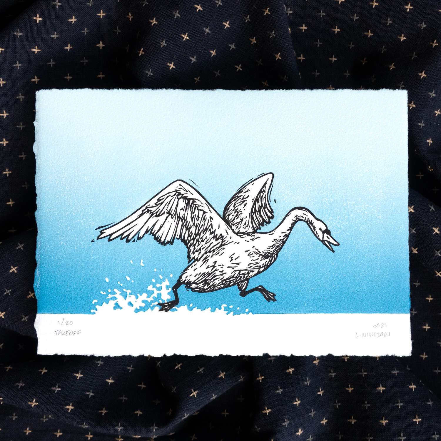 A 2-color print of a swan splashing on the surface of a lake as it takes off. The swan's wings are spread and the water and water spray are depicted using the uninked white paper. The sky is a blue-to-light blue gradient. The edges of the print are fuzzy and the print sits above a dark rumpled piece of fabric.