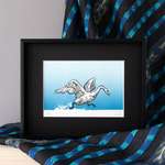 A graphic print of a swan taking off from the surface of the lake, framed behind a black mat in a thin black metal frame. The frame stands upright on a wooden shelf amongst the folds of a black and blue cascade of fabric.