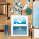 Top-down view of a desk, with the blue gradient layer printed on a rectangular paper at center. Below the print is the carved linoleum block for that layer, inked in a matching blue color. Printing tools and ink are scattered around.